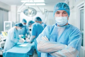 A surgical technician with arms crossed looking at the camera with a surgery taking place in the background.