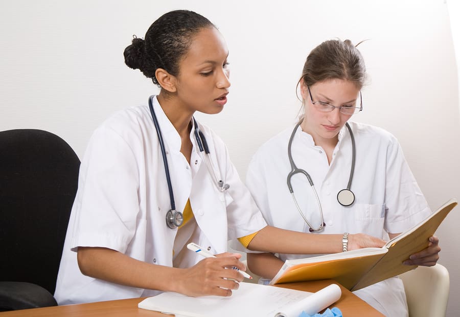 Two nurses sitting at a table looking at each others notebooks