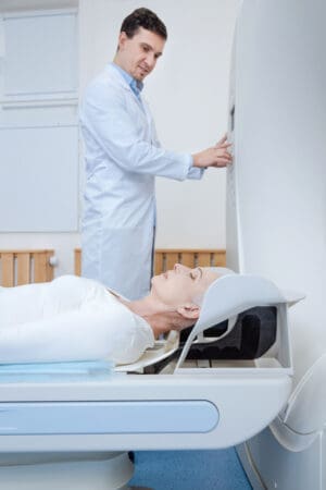 Elderly woman lying on an examination table and closing her eyes getting ready for an MRI while a technician works on the machine