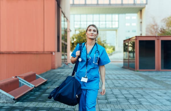 A nurse in scrubs, carrying a duffle bag and walking to a hospital.