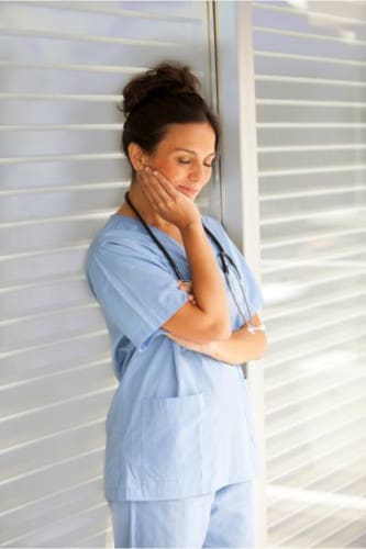 A nurse standing in front of a window with her hand under her chin thinking
