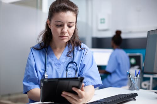 A nurse in scrubs reading a patient chart on a tablet.
