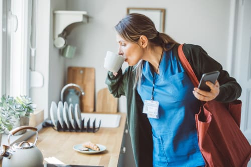 A nurse standing over a sink, drinking coffee, looking at her phone, and trying to eat breakfast