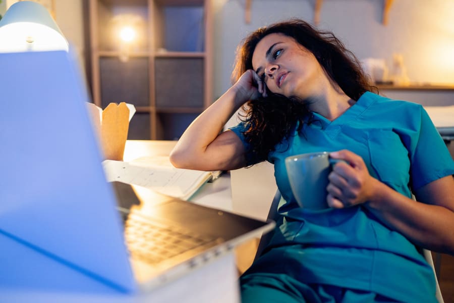 A nurse sitting at a desk holding a cup of coffee and looking tired.